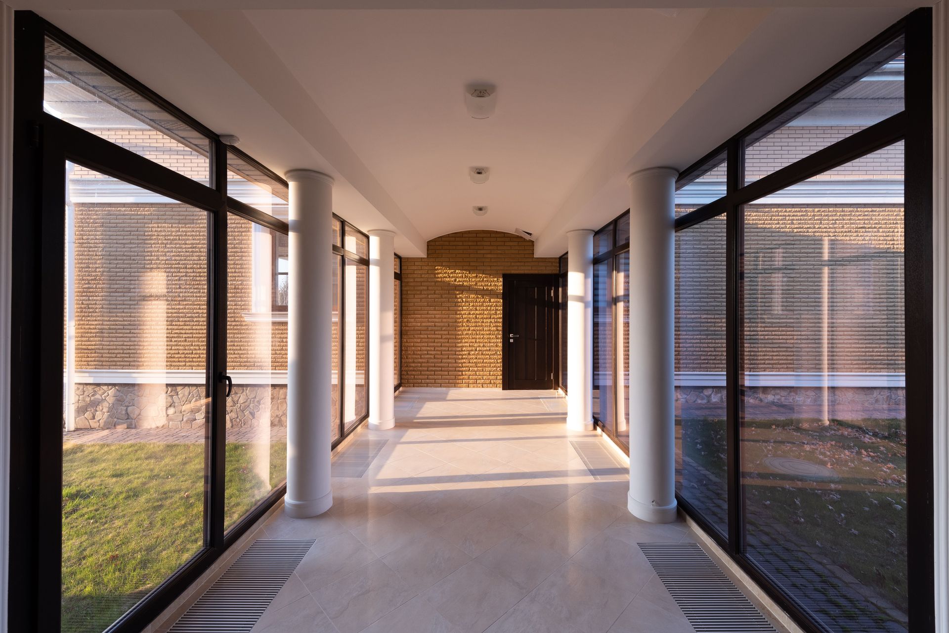 Empty long hallway with columns leading to brick wall and door between glass transparent walls of modern building overlooking grassy lawn